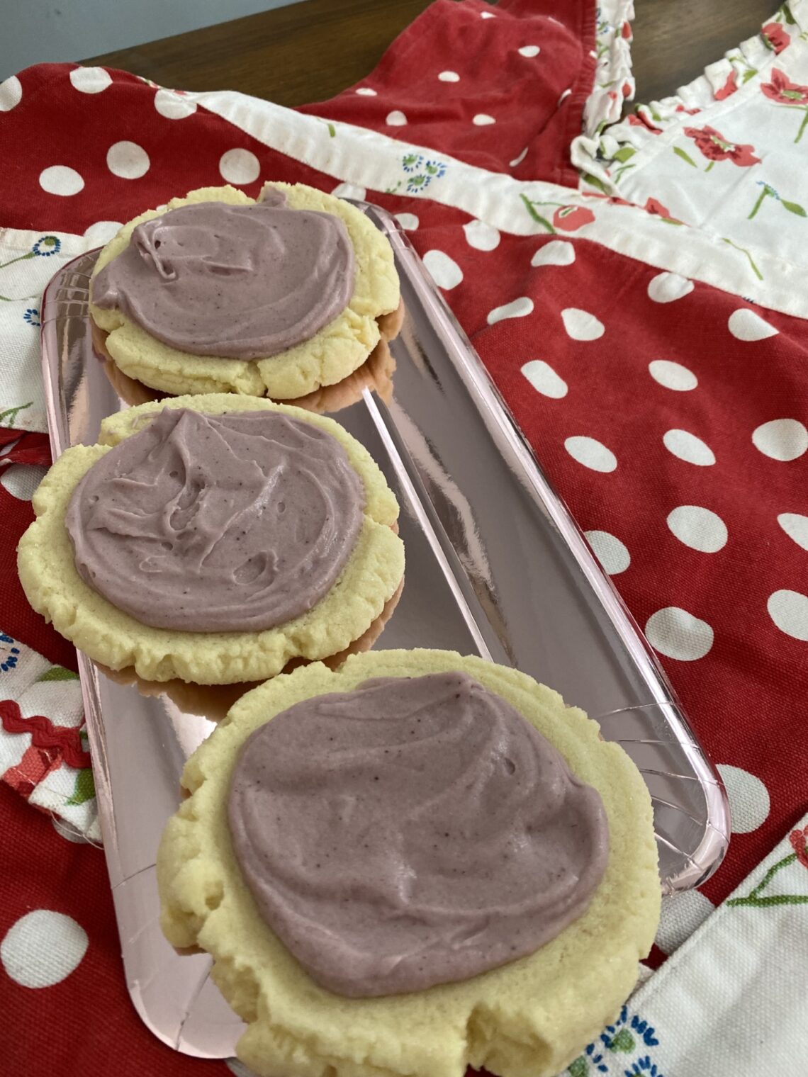 Picture featuring 3 sugar cookies with light purple frosting. The cookies sit on a foil plate atop a red and white polka dotted apron.