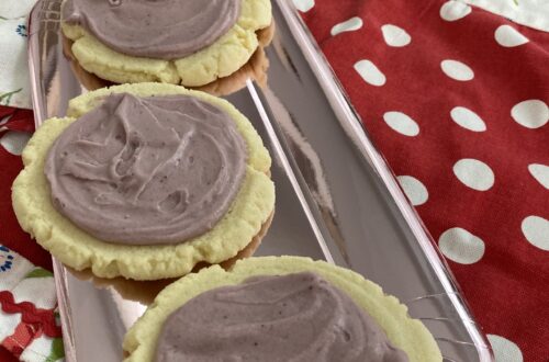 Picture featuring 3 sugar cookies with light purple frosting. The cookies sit on a foil plate atop a red and white polka dotted apron.