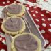 Picture featuring 3 sugar cookies with light purple frosting. The cookies sit on a foil plate atop a red and white polka dotted apron.