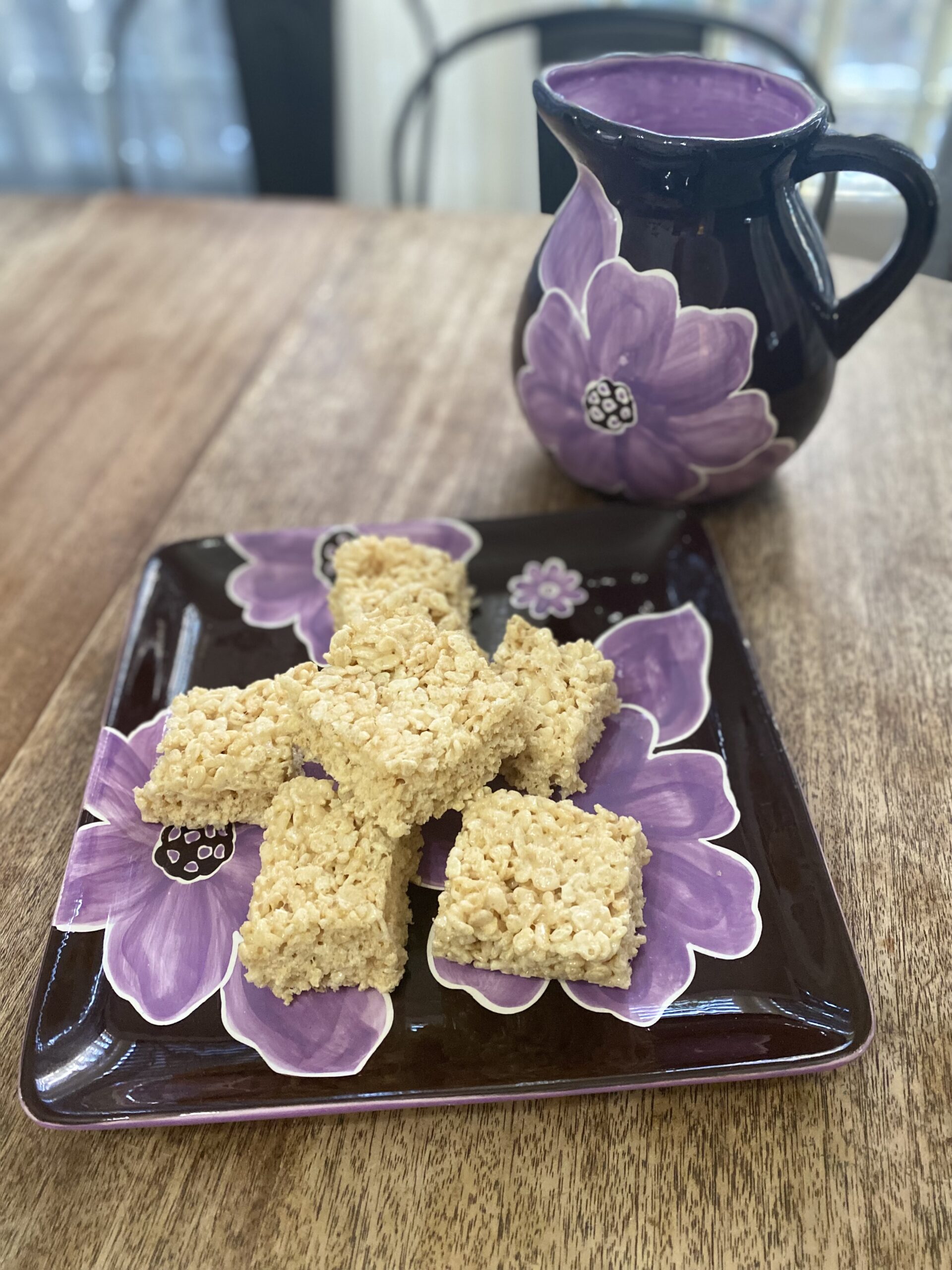 Marshmallow Crispy Treats sitting on a summery, colorful platter with a pitcher behind the platter.