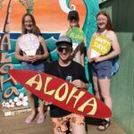 A dad and his kids are holding snowcones and a surfboard in front of a mural of a surfing wave.