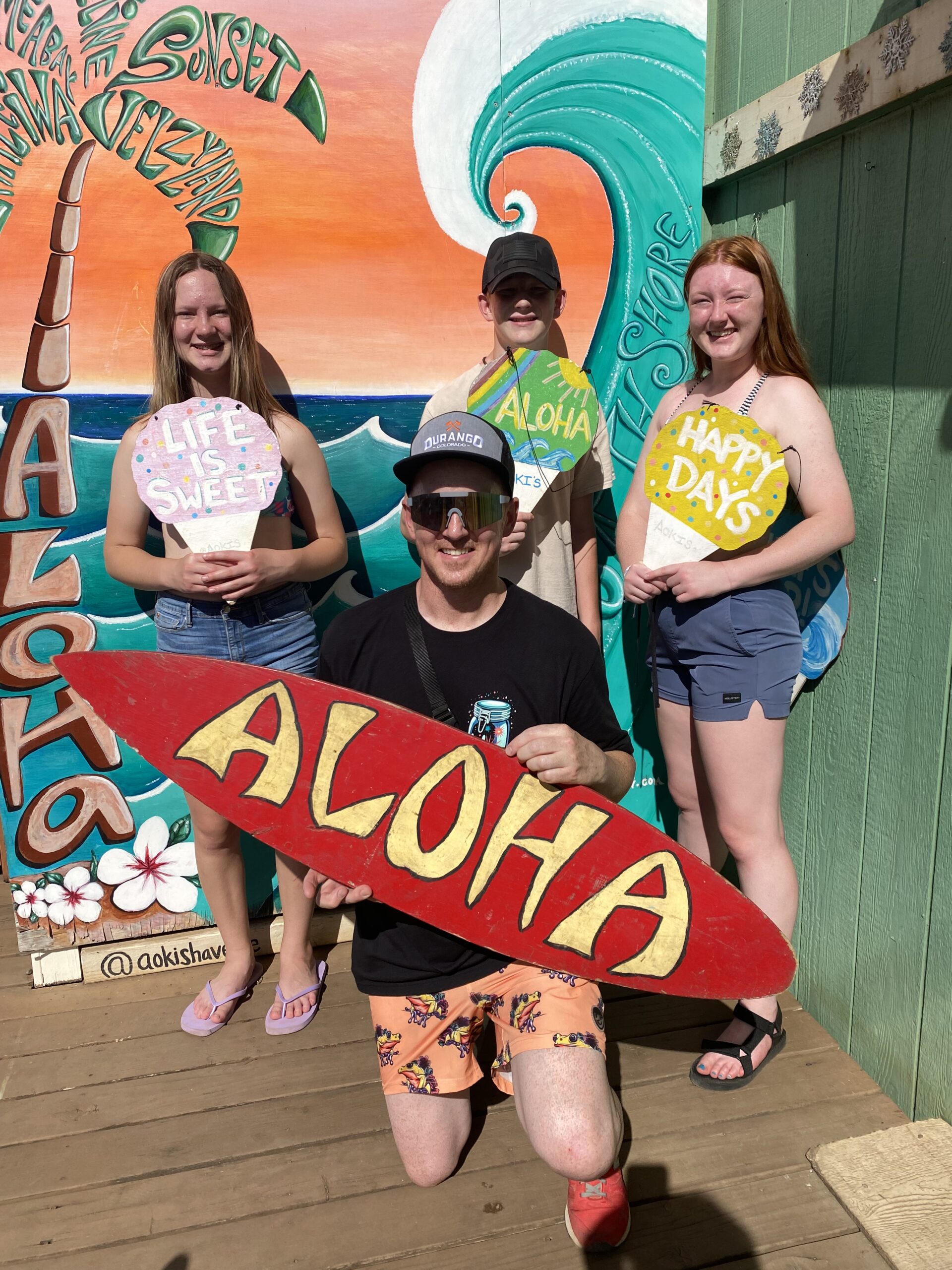 A dad and his kids are holding snowcones and a surfboard in front of a mural of a surfing wave.