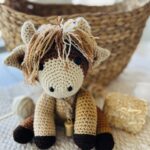 Crocheted Baby Highland Cow is sitting in front of a basket and a small hay bale.