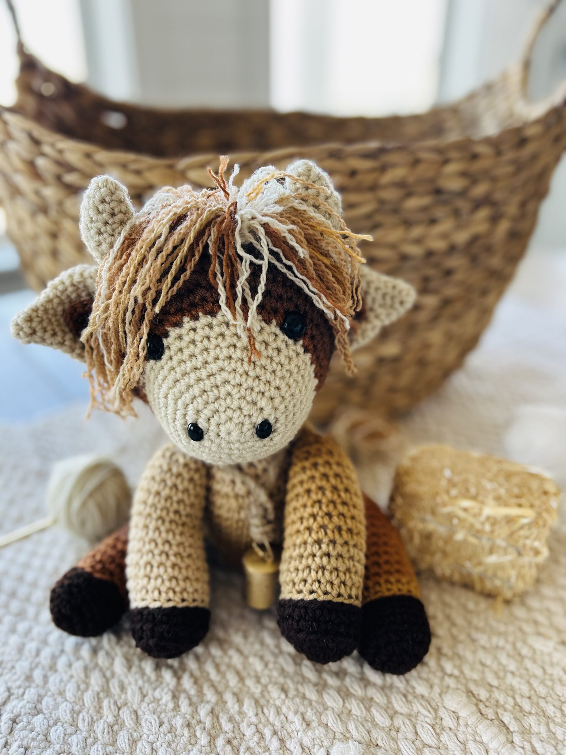 Crocheted Baby Highland Cow is sitting in front of a basket and a small hay bale.