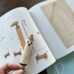 A Tan Crocheted Crayon is being held in front of a picture in a Childrens Book.