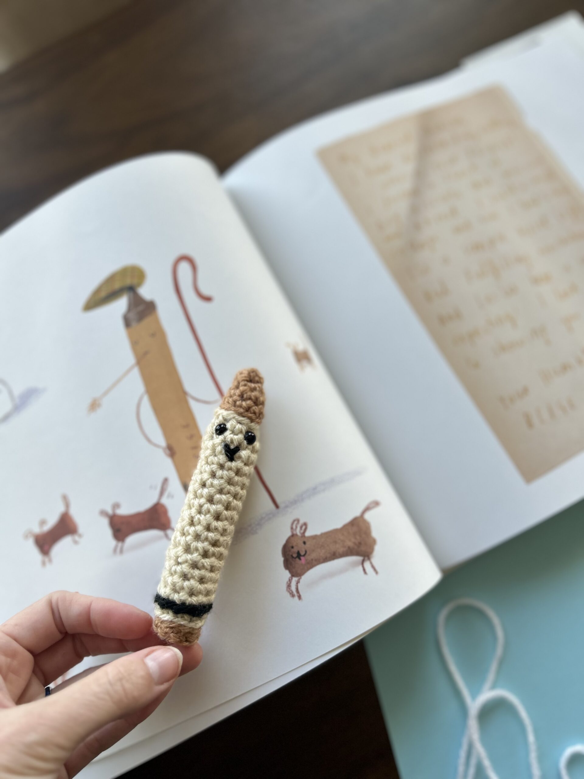 A Tan Crocheted Crayon is being held in front of a picture in a Childrens Book.